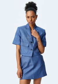 Blauwe blazer met korte mouwen en bijpassende minirok, gemaakt van textuurstof. De blazer heeft twee voorkeurszakken en een knoopsluiting.