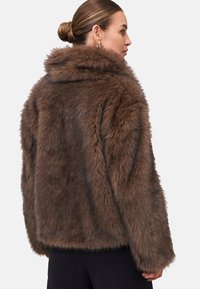 Veste en fausse fourrure marron avec un col haut, coupe décontractée et texture prononcée. Elle présente de longues manches et des coutures latérales. Aucun système de fermeture visible.