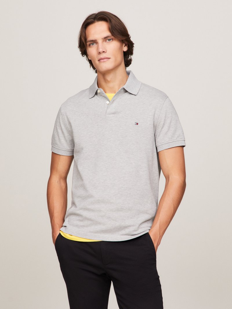 Grijze polo met korte mouwen en een kraag, met een subtiele logo op de borst; gedragen over een gele onderhemd; gecombineerd met zwarte broek.