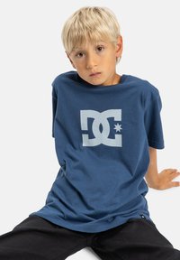 Navyblaue Baumwoll-T-Shirt mit einem großen weißen DC-Logo auf der Vorderseite. Verfügt über eine lockere Passform und kurze Ärmel, kombiniert mit schwarzen Hosen.
