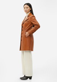 Brun trenchcoat i läder med figursydd design, slitsad krage och sex knappar. Kombinerad med vita vida byxor och svarta stövlar med klack.