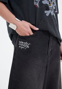 Schwarze Denim-Hosen mit einer Fronttasche und weißen, gestickten "Don Ed Hardy designs". Kombiniert mit einem lockeren schwarzen Shirt und einem metallischen Armreif.