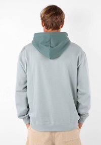 Haut à capuche gris clair avec une capuche gris foncé. Fabriqué en tissu doux, il présente des poignets et une taille côtelés, avec un détail de logo sur l'épaule.