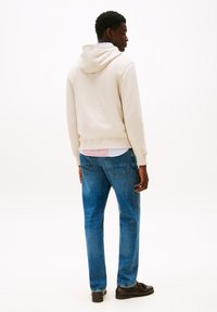 Man med beige hoodie, lager av skjortor, blå jeans och m�örkbruna loafers, som står och vänder sig åt sidan mot en vit bakgrund.