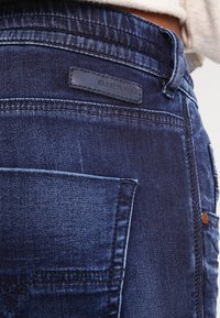 Vaqueros de denim en azul oscuro con una textura suave. Las características incluyen un bolsillo trasero, detalles de costura y un parche del logo "Diesel" visible.