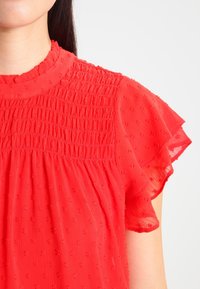 Czerwona, lekka bluzka z marszczonym stanem, krótkimi falbaniastymi rękawami i teksturowanym materiałem z małymi haftowanymi wzorami.
