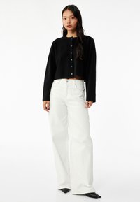 Schwarzer, kurzer Pullover mit Knopfverschluss, kombiniert mit weißen, weit geschnittenen Jeans und spitzen schwarzen Schuhen, die glatte Texturen und neutrale Farben aufweisen.