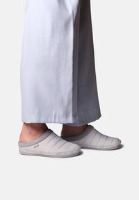 Pantoufles en cuir blanc matelassées avec des accents en tricot gris et une semelle douce et texturée, associées à un pantalon large gris clair.