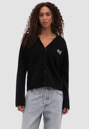 Kobieta o kręconych włosach, ubrana w czarny kardigan na guziki z białym haftowanym logo oraz jasnoniebieskie jeansy z wysokim stanem, stojąca.