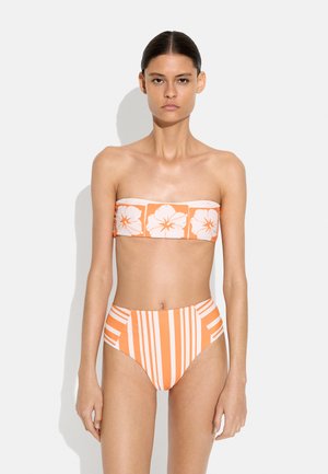 Kobieta w pomarańczowo-białym, bezramiączkowym topie bikini z kwiatowymi nadrukami i wysokim stanem w paski, stojąca na białym tle.