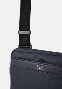 Bolsa de mensajero de tela azul marino con superficie texturizada, cierre con cremallera y una correa ajustable negra con un acento de logo cerca de la sujeción.