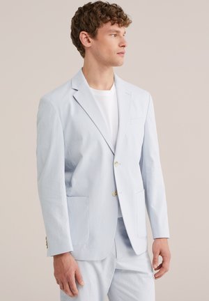 Jeune homme aux cheveux bouclés portant un costume bleu clair texturé et une chemise blanche, regardant vers sa droite sur un fond uni.