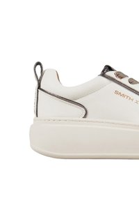 Sneaker bianca con suola spessa, parte superiore in materiale sintetico liscio, accenti metallici e linguetta sul tallone. Presenta lacci rotondi e dettaglio del logo.