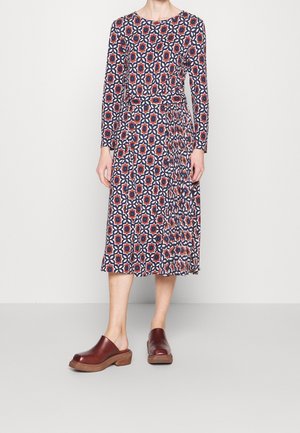 Robe à manches longues avec un motif géométrique en rouge, marine et blanc, présentant un haut ajusté et une jupe évasée plissée. Associée à des sabots marron.