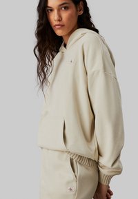 Sudadera beige hecha de tela suave, con un bolsillo frontal, puños acanalados y un pequeño parche del logo en el pecho. Corte casual y textura acogedora.