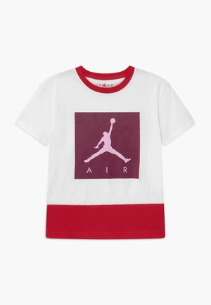 Camiseta blanca con cuello y dobladillo rojos, que presenta un gran cuadrado marrón rojizo con la silueta rosa de un jugador de baloncesto y el texto "AIR" debajo.