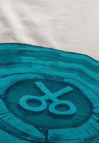 Stampa grafica su tessuto beige con forbici color turchese all'interno di un design circolare, che evidenzia linee spesse e un aspetto texturizzato.