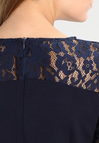 Robe bleu marine avec une délicate superposition de dentelle aux épaules, texturée de motifs floraux et une fermeture éclair discrète à l'arrière.