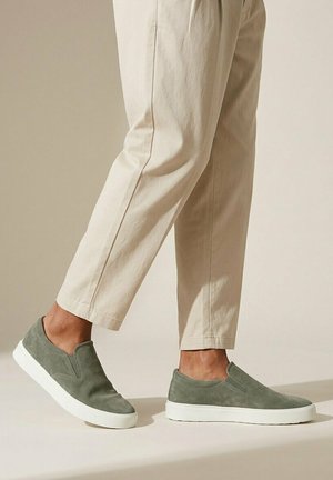 Benedenbenen met beige broek en olijfgroene instappers met witte zolen, staand op een neutrale achtergrond.