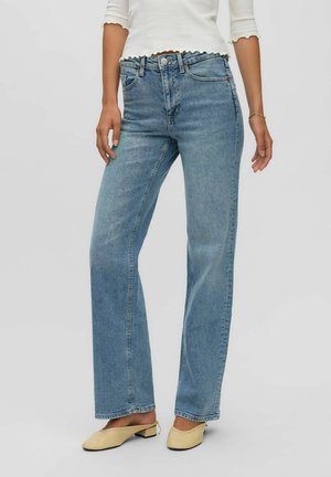 Femme portant un jean droit taille haute bleu clair, des chaussures crème à talons bas et un top blanc court à manches longues sur un fond uni.