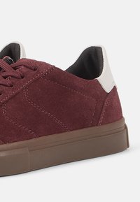 Maroon suede sneaker with a textured finish, featuring brown rubber sole, white heel tab, and brown laces. Simple, low-profile design.