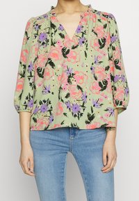 Femme portant un chemisier vert clair à motifs floraux roses, violets et noirs, et un jean bleu, debout devant un fond uni.