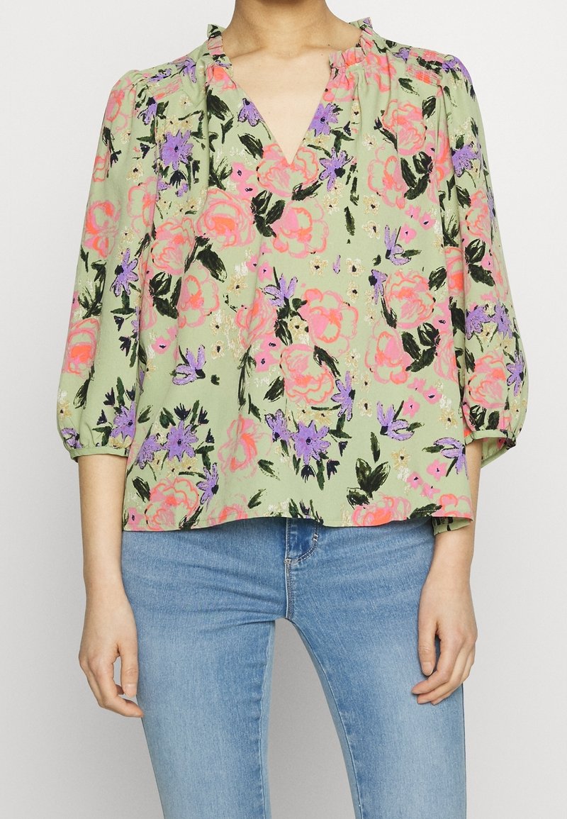 Femme portant un chemisier vert clair à motifs floraux roses, violets et noirs, et un jean bleu, debout devant un fond uni.