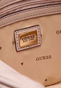 Interno in tessuto beige con testo stampato nero "GUESS" e un'etichetta in metallo dorato con la scritta "GUESS EST. 1981" cucita all'interno di una borsa.