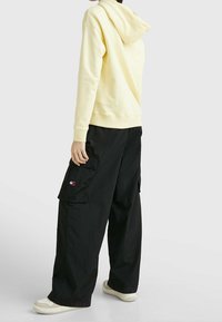 Sudadera amarilla con capucha y bolsillo tipo canguro, combinada con pantalones cargo negros de corte holgado que cuentan con bolsillos laterales. Zapatillas crema completan el look.