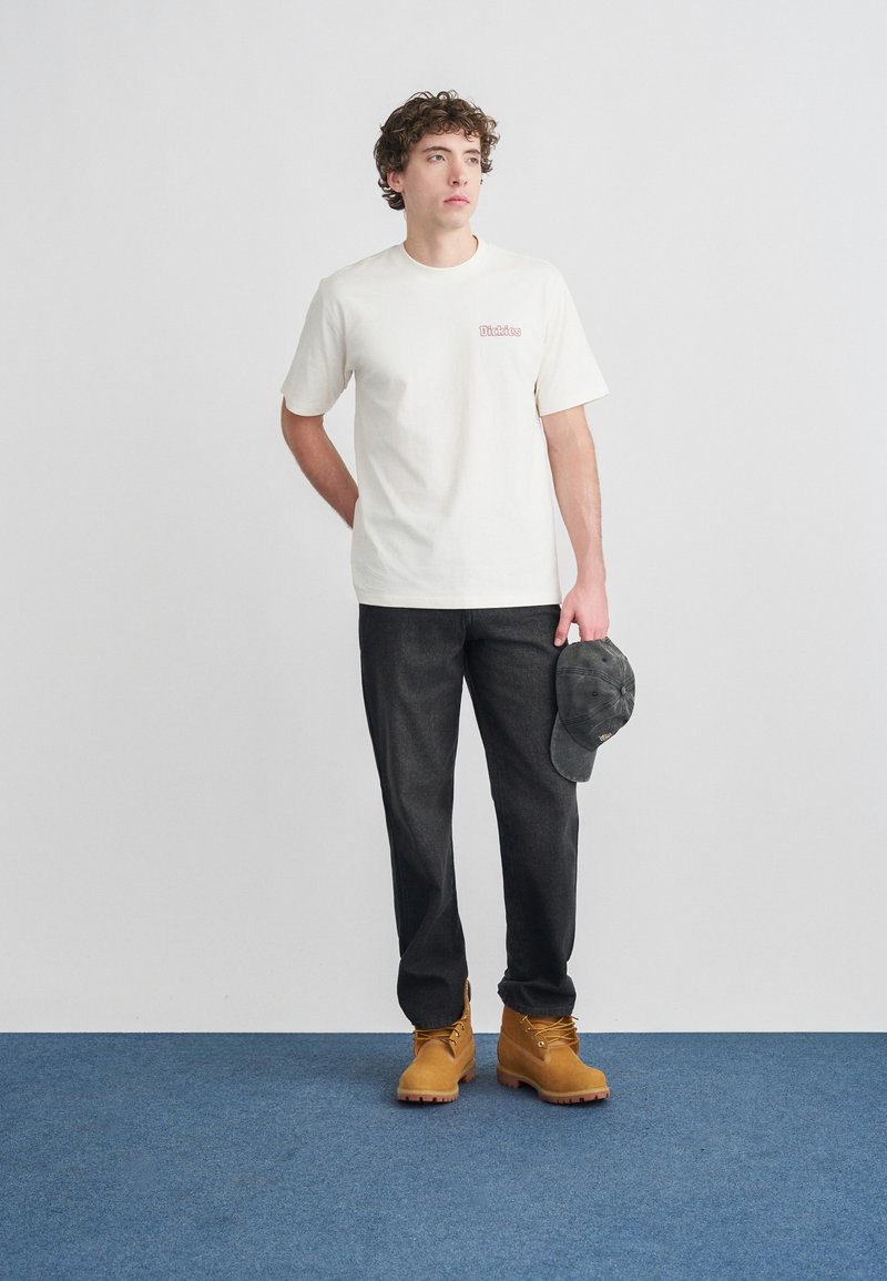 Hvid bomulds-T-shirt med rød logo, mørkegrå bukser og gulebrune arbejdssko. Holder en mørkegrå kasket. Simpelt, funktionelt sæt tøj.