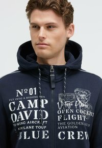 Sudadera con cremallera en color navy, confeccionada en tejido de mezcla de algodón, con texto impreso en blanco, capucha ajustable con cordón y detalles de costura en contraste.