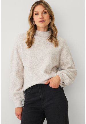 Kvinde med blondt hår iført en lysebeige strikket sweater og sorte højtaljede bukser, stående med den ene hånd i lommen mod en hvid væg.