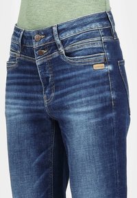 Dark blue denim jeans with a faded finish, two-button closure, and five-pocket design. Features subtle stitching and a small brand label.