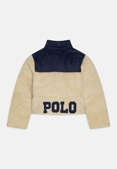 Rövidített dzseki bézs, bolyhos alsó résszel és sötétkék nylon felső résszel. A hátsó részen sötétkék hímezett "POLO" felirattal.