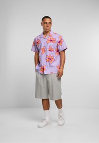 Lilla t-skjorte med korte ermer og stort oransje blomsterdesign, kombinert med lysegrå shorts og hvite sneakers. Enkel, uformell stil.