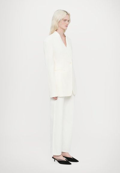 Witte op maat gemaakte blazer met een enkele knoop, brede revers, gecombineerd met witte wijde broek en zwarte puntige slingback hakken.