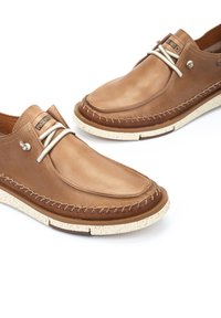Mocasines de cuero marrón con un diseño cosido, cordones gris claro y una suela de goma beige moteada. Incluye un detalle de marca minimalista.
