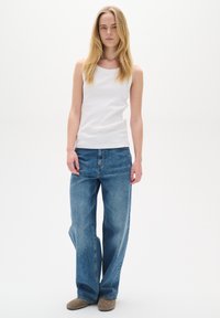 Jeune femme aux longs cheveux blonds portant un haut blanc sans manches, un jean bleu ample et des chaussures marron, debout devant un fond blanc.