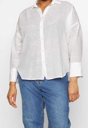 Person iført en løs hvid skjorte med brede manchetter og blå denimjeans, stående mod en ensfarvet baggrund.