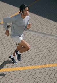 Světlě šedý dlouhý rukáv, béžové šortky a černé sportovní boty. Běžící postava na kamenném chodníku s žlutou čárou v pozadí.