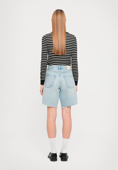 Shorts in denim azzurro con orli sfrangiati, abbinati a una camicia a righe bianco e nera a maniche lunghe, indossati con calzini bianchi e scarpe nere.