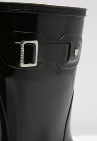 Botte en caoutchouc noir brillant avec un haut arrondi, dotée d'une boucle en métal argenté et d'un logo en relief. Texture lisse avec un design épuré et un minimum de détails.