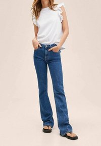 Witte mouwloze top met rimpeldetails, gecombineerd met een hoge taille, wijde blauwe jeans en zwarte sandalen. Eenvoudig ontwerp met een casual pasvorm.