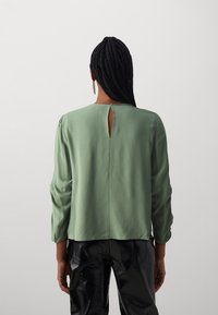 Grön blus med lång ärm och nyckelhålsdetalj på baksidan. Slät tyg, avslappnad passform och samlade ärmslut. Matchad med glänsande svarta byxor.