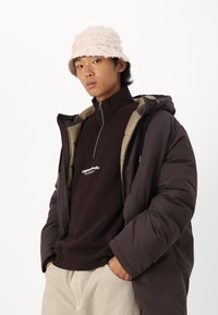 Chaqueta acolchada marrón con un forro color beige claro, cuello alto y cremallera. Llevada sobre un sudadera marrón oscuro con logo. Sombrero tipo bucket texturizado en rosa claro.
