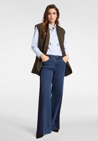 Olivově zelená bezrukávová vesta přes světle modrou košili s květinovou výšivkou, v kombinaci s širokými tmavě modrými džíny a hnědými podpatky.