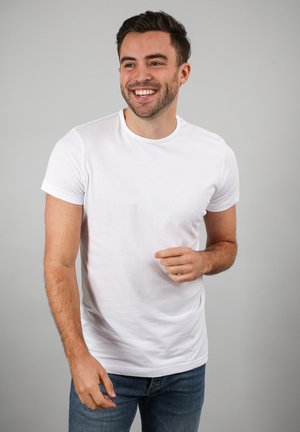 Glunderende man met kort bruin haar, die een simpel wit T-shirt en blauwe spijkerbroek draagt, staand tegen een lichtgrijze achtergrond.