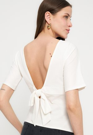 Nő fehér, rövid ujjú, nyitott hátú, masnival megkötött felsőben, sötét farmernadrággal és arany karika fülbevalóval.