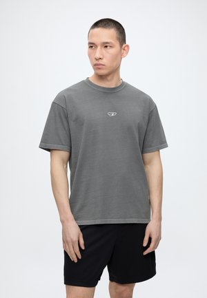 Młody mężczyzna z krótkimi włosami w szarej, luźnej koszulce z małym logo i czarnych szortach, stoi na tle jednolitego jasnego tła.