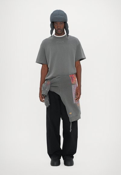 T-shirt gris à manches courtes avec des détails de couture, associé à un pantalon noir ample. Un hoodie gris détachable tombe à la taille, arborant des accents de motifs colorés.